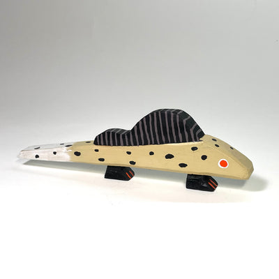 Painted whittled wood sculpture of a simplistic lizard, tan with black polka dots and a dorsal fin. It has small feet and a red eye.