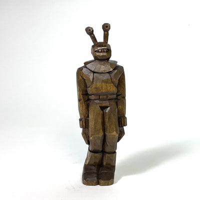 Stained whittled wood sculpture of an alien with eyes like a snail, wearing a full space suit with boots and standing awkwardly with slightly bent knees.