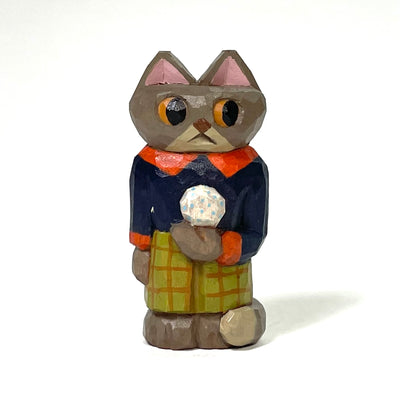 Painted whittled wood sculpture of a brown cat, standing on 2 legs with its hands to its side. It wears a collared shirt, plaid pants and holds a snowball.