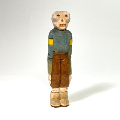 Painted whittled wood sculpture of a skeleton with small, sunken red eyes and yellow teeth. It stands with its long arms at its side, wearing a blue sweater and tan pants.