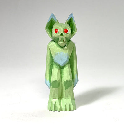 Painted whittled wood sculpture of a green goblin with a cute face, red eyes and pointed ears. It stands with its hands at its side, looking off curiously.