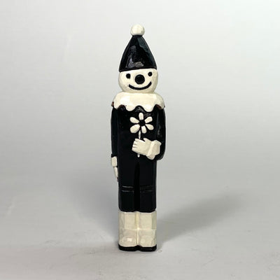 Painted whittled wood sculpture of a clown, black and white with an old school cartoon look to it. It stands tall, smiling and holding a single white flower.