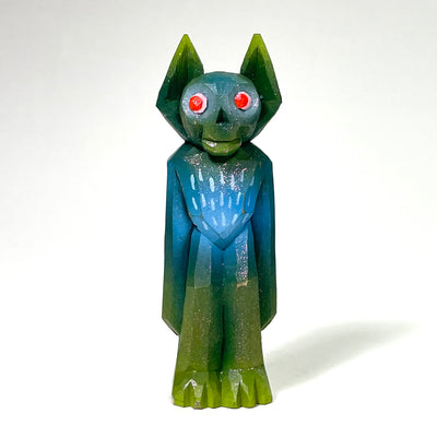 Painted whittled wood sculpture of a blue and green goblin with a cute face, red eyes and pointed ears. It stands with its hands at its side, looking off curiously with a smile.