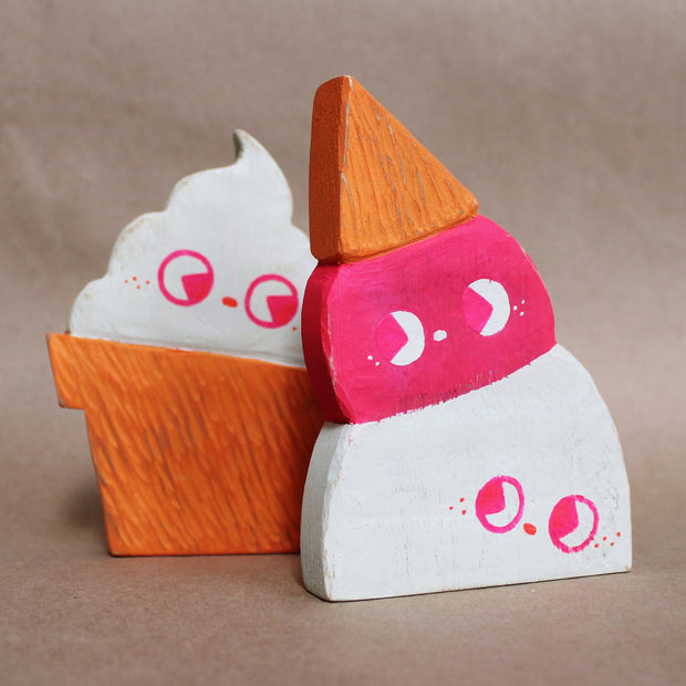 Die cut painted wooden sculpture of an upside down double scoop ice cream cone. One scoop is pink and the other is white, they both have cute drawn on faces looking off to the side. It stands in front of a die cut painted wooden sculpture of a soft serve cone.