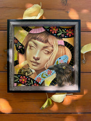 Finely rendered colored pencil illustration of a woman, drawn in sepia coloring. She looks straight on to the viewer. Around her are crows and 2D floral patterned snakes. Piece is framed in a wooden frame without glass.