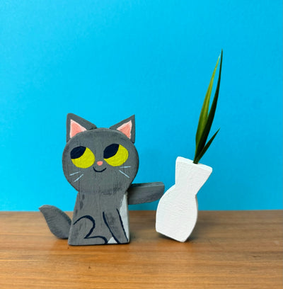 Wooden sculpture of a cute gray cartoon cat, with yellow eyes looking mischievously off to the side. It begins to push over a wooden vase with a plant in it.