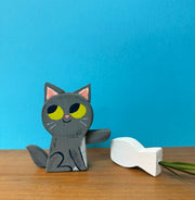 Wooden sculpture of a cute gray cartoon cat, with yellow eyes looking mischievously off to the side. It pushes over a wooden vase with a plant in it.