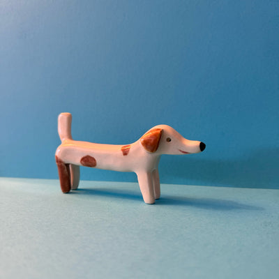 Small ceramic sculpture of a long white dog with brown spots. It has simplistic facial features and a simple smile.