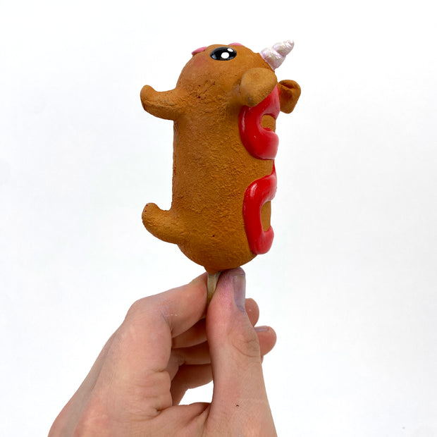 Sculpture of a corn dog, designed to look like a dog with a cute, silly face and a metallic unicorn horn atop its head. It has a squiggle of ketchup on its back.