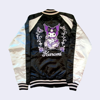 Shiny polyester bomber jacket with black body and white sleeves. Back has a large embroidered graphic of Kuromi sitting on a purple rose with more roses and butterflies next to her.