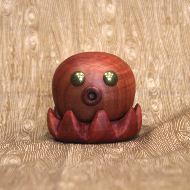 Wooden sculpture of a small octopus, with shiny metal eyes and an "o" shaped mouth. Its tentacles curl up towards itself.