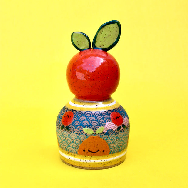 Ceramic sculpture of a bright colored orange, acting as a knob handle atop of an upside down dome shape. The dome has paintings of semi circle patterns and other oranges.