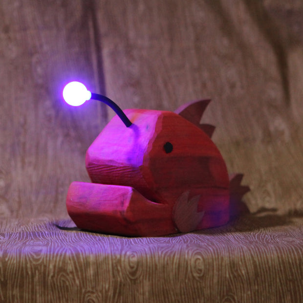 Whittled red wooden sculpture of an angler fish, with large cartoon type proportions. It has a light coming out of its head, emitting a purplish glow.