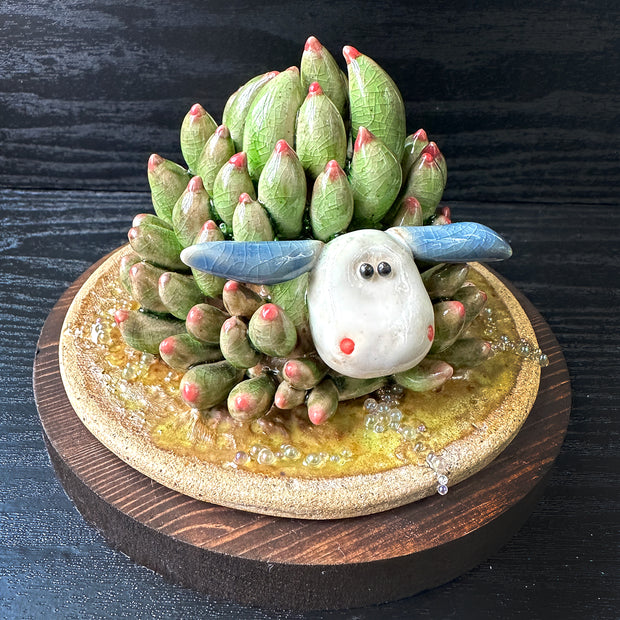 Sculpture of a white and blue sea slug with its back akin to a succulent, green with pink tips. It lays on a round ceramic base that has a green watery sheen. 