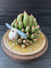 Sculpture of a white and blue sea slug with its back akin to a succulent, green with pink tips. It lays on a round ceramic base that has a green watery sheen.