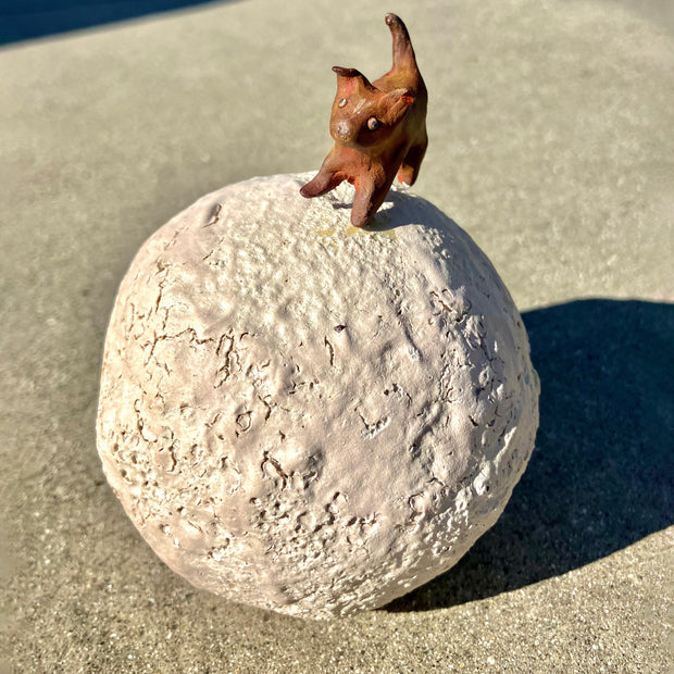 Ceramic sculpture of a round crater like moon or planet, with a small brown dog standing on top of it.