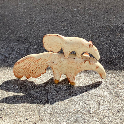 Cream colored ceramic sculpture of a large anteater walking with a slightly smaller anteater perched atop its back. Both have speckled glazes and slight rust brown detail coloring.