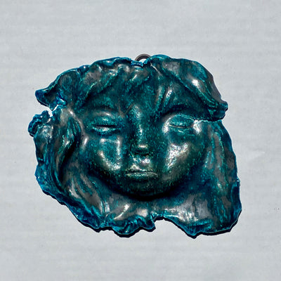 Blueish sculpture of a person's face, mostly flat with their hair flowing around it. Their eyes and mouth are closed.