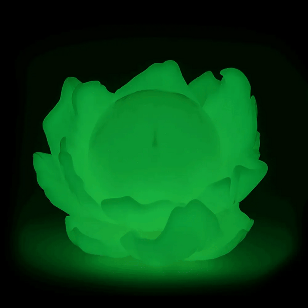 Glow in the dark colored vinyl figure of a round full moon with a small minimalistic face. It sits within a series of flower petals, as if it is the pistil of the flower. Gif goes from lights off, to show glow, to lights on.