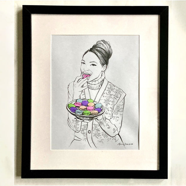 Illustration on white paper of a girl with her hair up in a fancy bun. She smiles and eats from a plate of macarons. Piece is framed.
