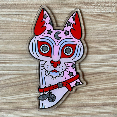 Die cut flat acrylic sheet, shaped like a Sphynx cat head. It wears a collar with a bell and fish charm and has stars spotting its skin. Its painted light pink and purple with red and silver color accents.