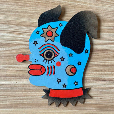 Painting on a die cut piece of wood of a cartoonish blue dog with a red clown nose and matching red lips. It has stars on its face like freckles.