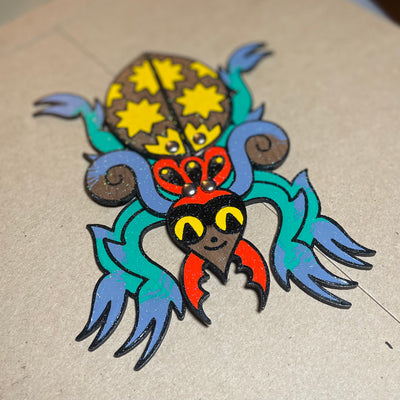 Die cut sculpture of a colorful spider, done in a tattoo art style with bold black lines and solid colors. It has a star patterned body, blue green legs and red pincers coming out of a smiling face. 