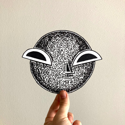 Ink drawing on a cut out circle of paper, with a moon texture and pair of semi circle eyes, glaring at the viewer. It has a straight mouth and serious expression.