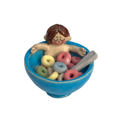 Ceramic sculpture of a nude girl, smiling and sitting inside a blue cereal bowl with Fruit Loops around her. She has a peaceful, closed eye expression.