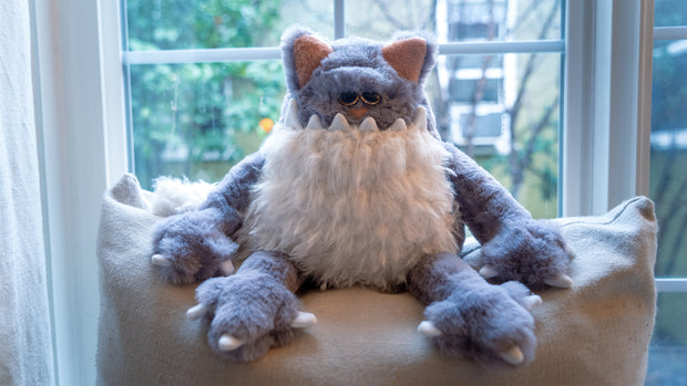 Fluffy plush sculpture of a gray cat, with a large jagged tooth underbite and a white fluffy belly that looks like a beard. It sits and looks off into the distance.