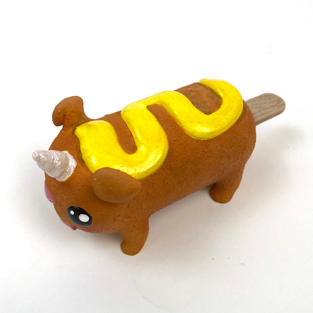 Sculpture of a corn dog, designed to look like a dog with a cute, silly face and a metallic unicorn horn atop its head. It has a squiggle of mustard on its back.