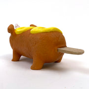 Sculpture of a corn dog, designed to look like a dog with a cute, silly face and a metallic unicorn horn atop its head. It has a squiggle of mustard on its back.