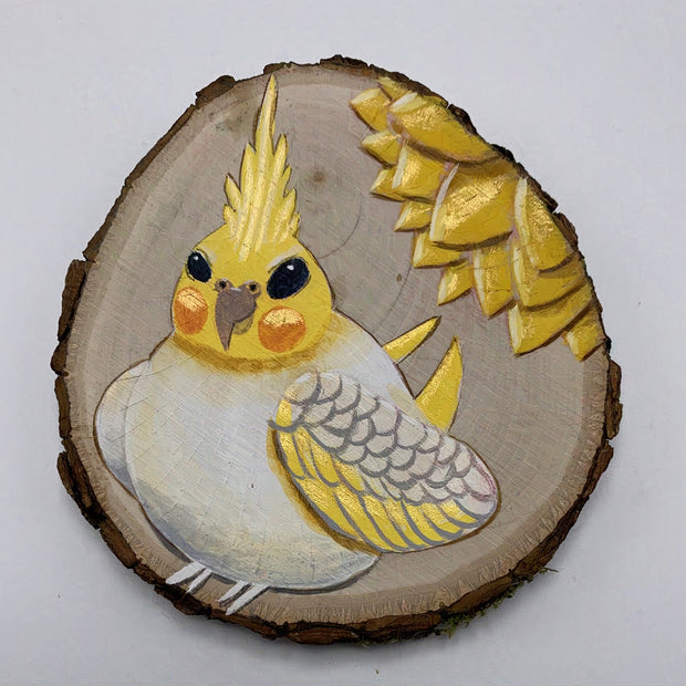 Painting on a circular slice of wood, with much of the wood grain left exposed. A cartoon style yellow cockatiel sits with a rotund and stout body. Behind it is a yellow flower, on its petals showing.