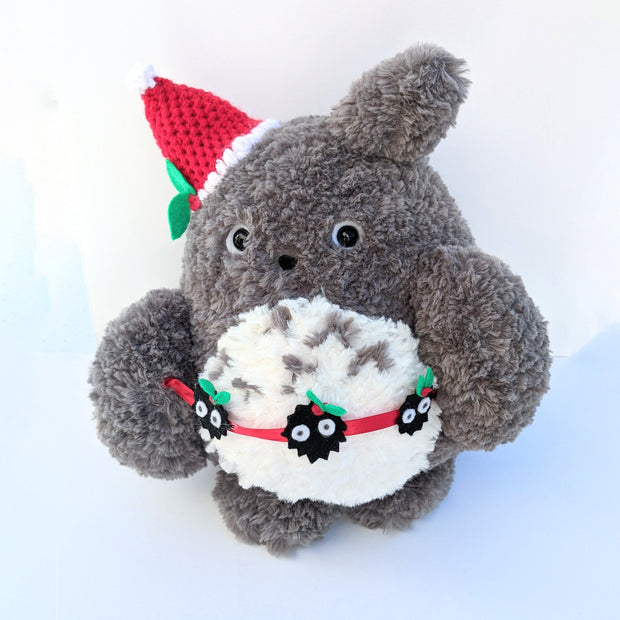 Rounded plush Totoro with a red crocheted Santa hat atop one of its ears. It holds a garland made of red ribbon and small black soot sprites with green sprouts on their heads.