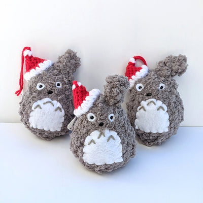 Group of small crocheted Totoros, all with red Santa hats hanging on one of their ears. 