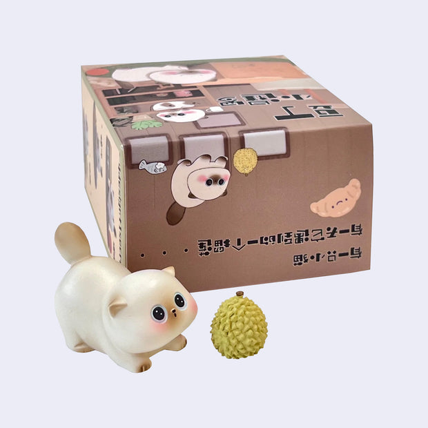 Small Siamese cat style figurine, with a large head and eyes. It plays with a durian and stands next to its product packaging.