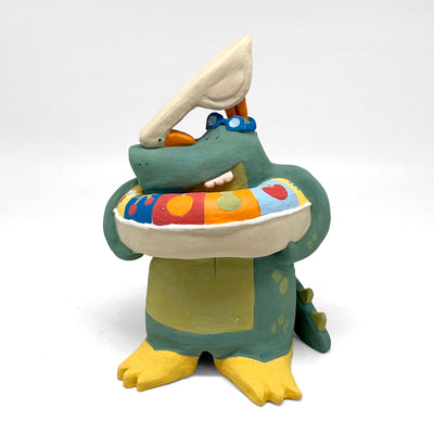 Ceramic sculpture of a green alligator standing up and wearing yellow flippers, goggles and with a inflatable tube around its belly. Atop its head is a white bird, looking down at the gator.