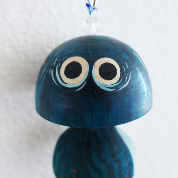 Sculptural wood piece, strung together so each piece hangs in a vertical row. Shapes are half domes, mostly painted blue with subtle patterns and details. The top dome has a set of large cartoon eyes.