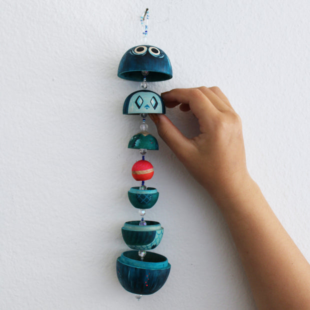 Sculptural wood piece, strung together so each piece hangs in a vertical row. Shapes are half domes, mostly painted blue with subtle patterns and details. The top dome has a set of large cartoon eyes.