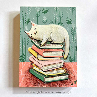 Painting of a white cat, resting atop a pile of books. The books are on a pink tablecloth with a mint colored background.