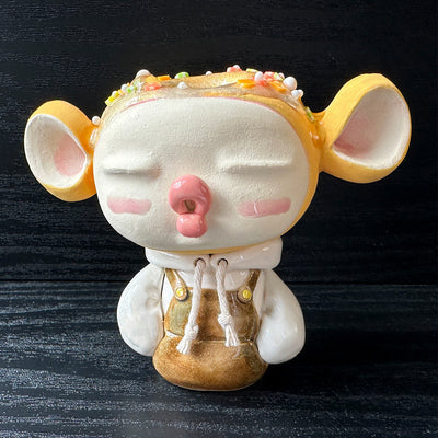Ceramic sculpture of a large headed character, with closed eyes and full lips, making a kissy face. They have big ears and wear a hoodie, with arms tucked in. Their head is orange with sprinkles atop, like a donut.