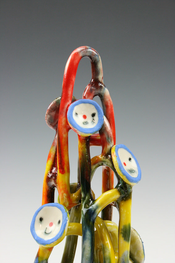 Ceramic sculpture of many looping shapes, like a stretched out pile of yarn. it is multicolor and has simple smiley faces like buttons, with blue outlines and simple features.