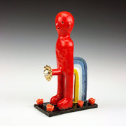 Sculptural piece of a red character with no arms and simple facial features and no arms. It stands and a flower comes out of its groin. Its propped up by a partial rainbow of blue, white and yellow. Roses bloom out of the base. 