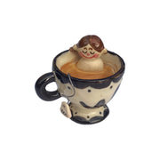 Small ceramic sculpture of a nude girl sitting inside of a tea cup surrounded by coffee colored liquid. A curled wire comes out of the mug with a small tag that reads "tea."