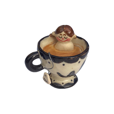 Small ceramic sculpture of a nude girl sitting inside of a tea cup surrounded by coffee colored liquid. A curled wire comes out of the mug with a small tag that reads "tea."