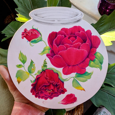 A hand holds an 8.5" flat illustration of a terrarium with three dark red roses inside. The roses are at varying stages of life. There is a bud, a full open rose and a downturned, old rose. The leaves on the older rose are slightly eaten and browning.