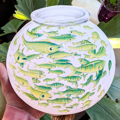 A hand holds an 8.5" illustration of a spherical fishbowl. There are over 60 fish of varying sizes, circling within the tight space of the fish bowl. They all have different subtle expressions varying from perturbed to joyful to confused.