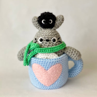 Crochet sculpture of a blue mug with a pink heart. Inside the mug is Totoro with a green scarf. A dust sprite sits atop its head.