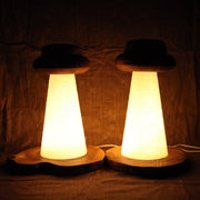 2 wooden lamps, both with UFO shaped tops and conical shaped beams, where light shines through. One lamp has a bean shaped wooden base, the other has a circular wooden base.