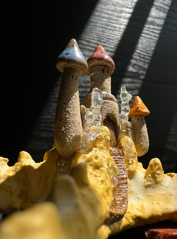 Ceramic sculpture of a yellow sea star with a sand colored castle atop of it, with multicolor pointed tower roofs. The sculpture sits atop a natural piece of wood.